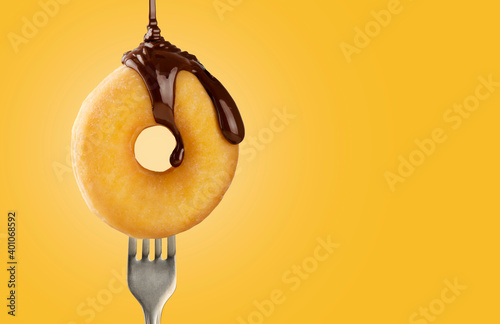 Topping of melted chocolate falling on a delicious doughnut that is pricked on a fork on a yellow background with copy space