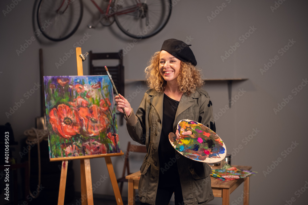 Fototapeta premium Young happy smiling female artist in an art workshop. Draws a beautiful abstract picture with flowers