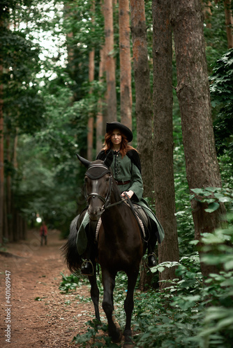 Young beautiful woman in a suit, green dress, corset, black topm hat on a black horse on nature in the forest. fairy tale, creative photo session of a girl with a horse