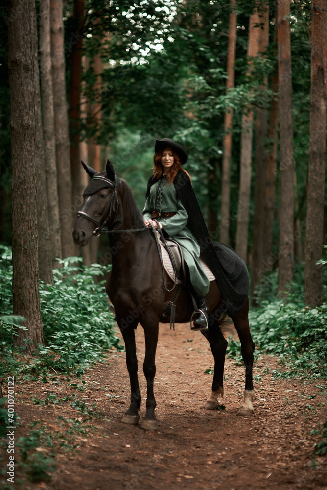 Young beautiful woman in a suit, green dress, corset, black topm hat on a black horse on nature in the forest. fairy tale, creative photo session of a girl with a horse Young beautiful woman in a suit, green dress, corset, black topm hat on a black horse on nature in the forest. fairy tale, creative photo session of a girl with a horse