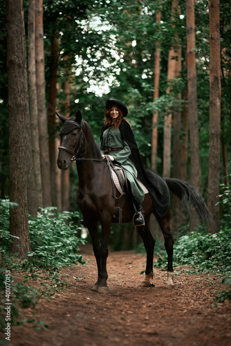 Young beautiful woman in a suit  green dress  corset  black topm hat on a black horse on nature in the forest. fairy tale  creative photo session of a girl with a horse
