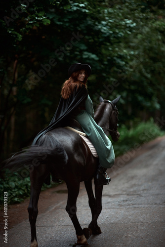 Young beautiful woman in a suit, green dress, corset, black topm hat on a black horse on nature in the forest. fairy tale, creative photo session of a girl with a horse photo