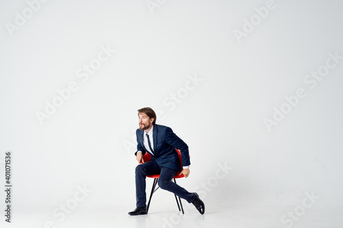 business man sits on red chairs indoors and gestures with his hands on a light background
