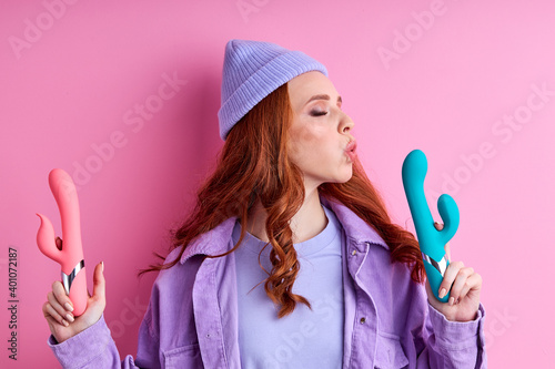 woman enjoys sexual life poses with vibrators to satisfy all types of preferences, have fun kissing it posing at camera isolated on pink background