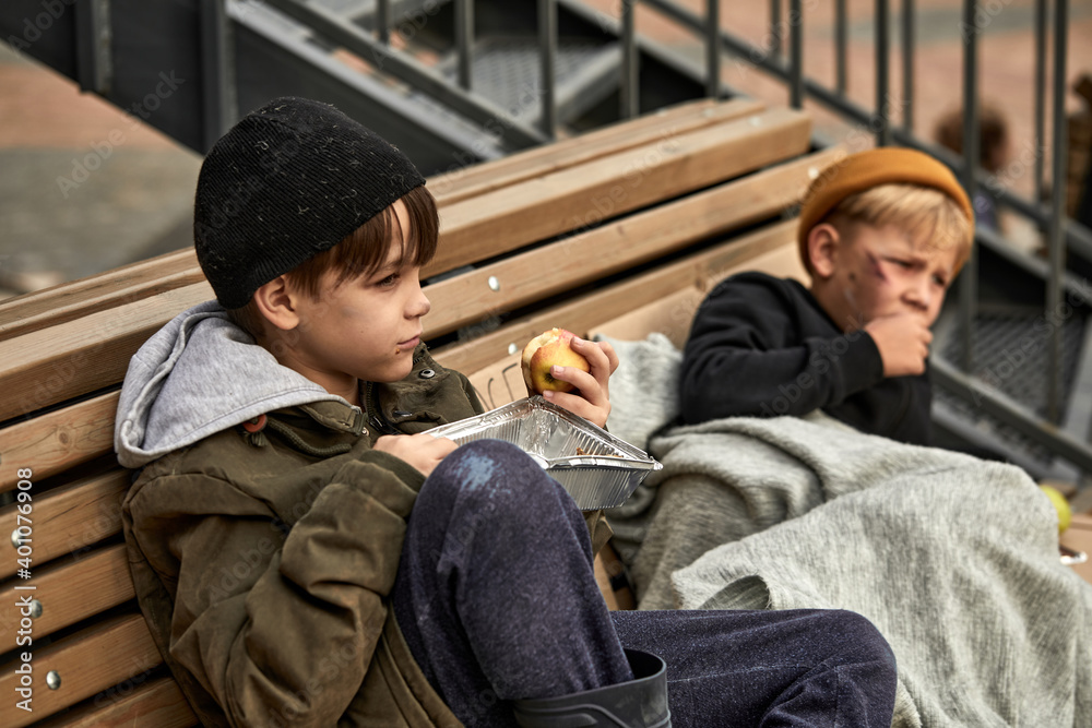 two homeless boys sit eating food on the bench, little caucasian children, street kids are hungry, get some food by kind people