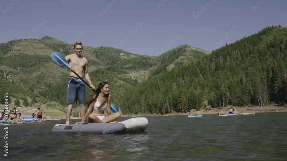 Wide shot to close up of approaching boy paddling girlfriend on paddleboard in lake / Tibble Fork, Utah, United States Wide shot to close up of approaching boy paddling girlfriend on paddleboard in lake / Tibble Fork, Utah, United States