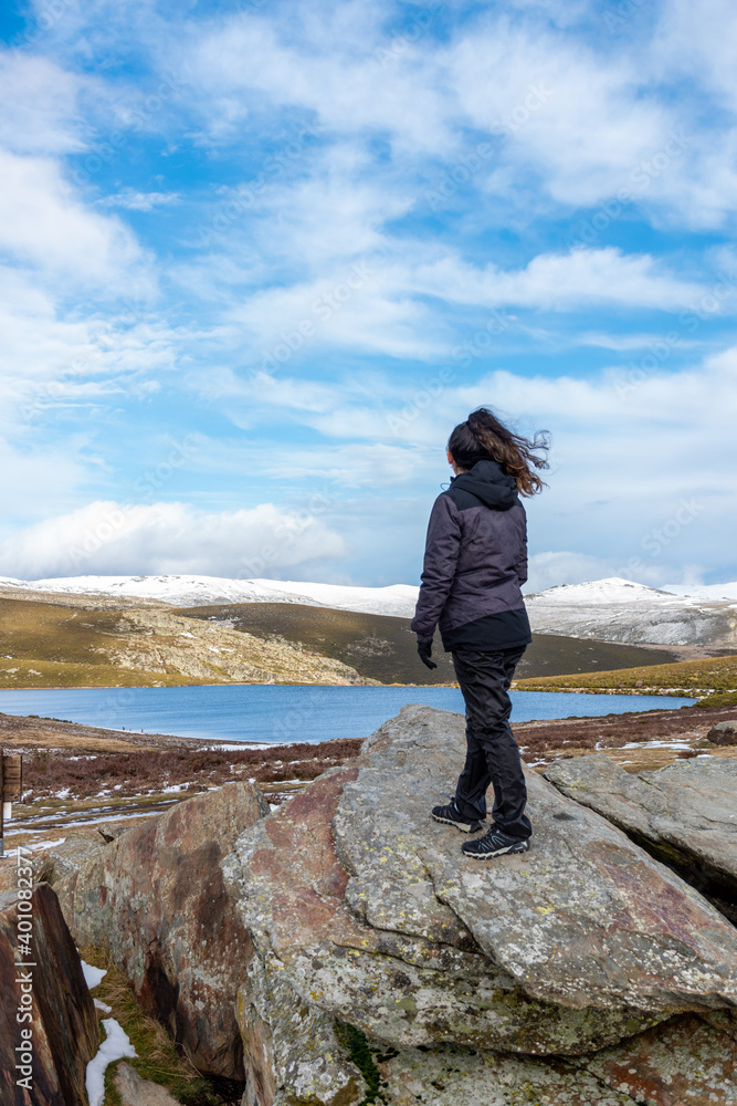 Obraz premium Vertical view of a white Caucasian woman on a rock overlooking a landscape of a lake and the snowy mountains in the background on a sunny day
