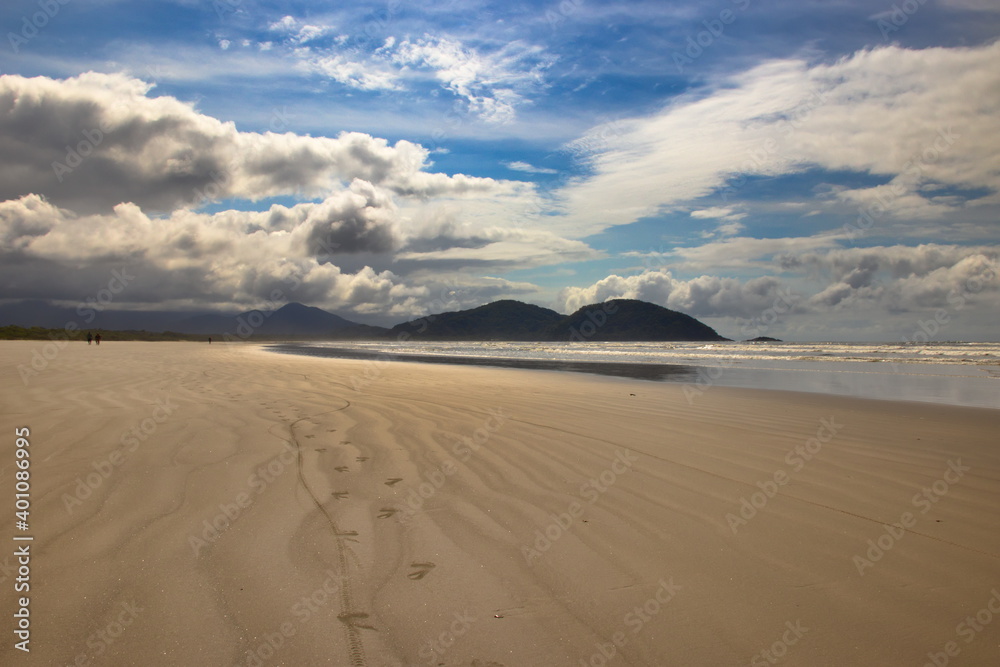 footprints heading towards the horizon, imprinted on the golden sands of the sparkling water beach, with mountains in the background under a blue sky with large white clouds.