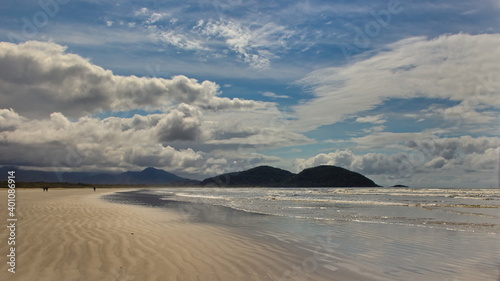 panorama image of a beach with a strip of golden sand waved by the tide, and a sparkling sea with small waves, in the background the distant mountains under a blue sky with large white clouds.