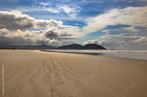 footprints heading towards the horizon  imprinted on the golden sands of the sparkling water beach  with mountains in the background under a blue sky with large white clouds.