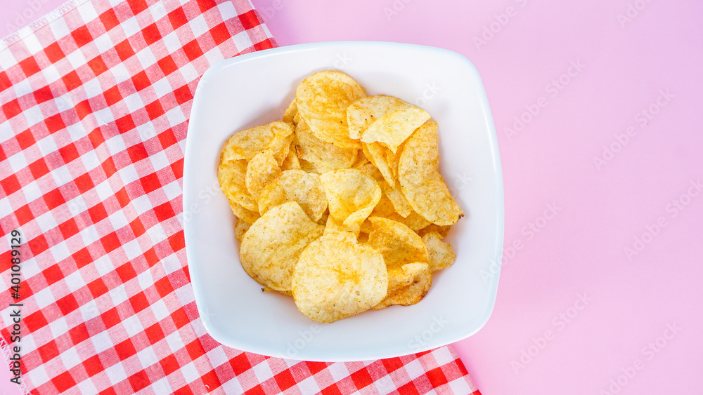 Naklejka premium Potato chips in bowl on napkin and pink background, top view. Salty crisps scattered on a table