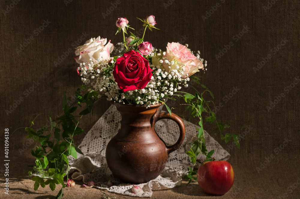 Still life in dark colors, with a bouquet of roses and gypsophila in a jug and a red Apple, on a background of burlap. Selective focus
