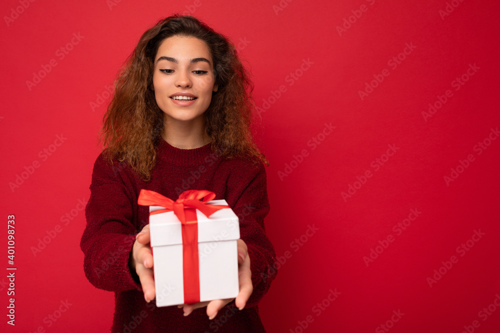 Attractive positive smiling young brunette curly woman isolated over red background wall wearing red sweater holding gift box looking at present white box with red ribbon Attractive positive smiling young brunette curly woman isolated over red background wall wearing red sweater holding gift box looking at present white box with red ribbon