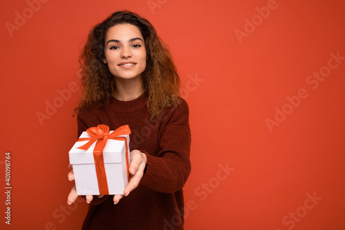 Beautiful happy young brunette woman isolated over colourful background wall wearing stylish casual clothes holding gift box and looking at camera © Ivan Traimak