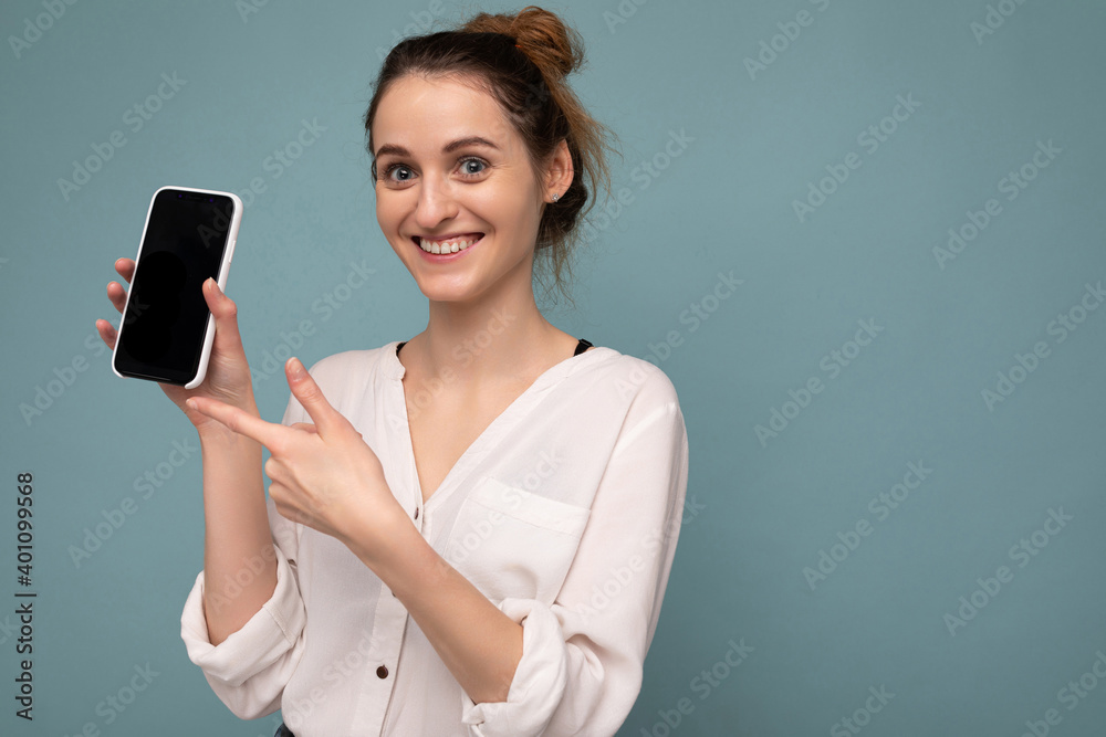Naklejka premium Beautiful positive young blonde curly woman wearing casual white shirt and isolated over blue background wall holding phone and showing smartphone with empty dcreen for mockup looking at camera and