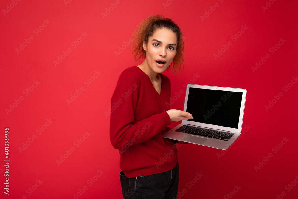Close-up portrait of beautiful amazed surprised astonished dark blond woman holding laptop computer looking at camera showing at netbook keyboard wearing red sweater isolated over red wall background Close-up portrait of beautiful amazed surprised astonished dark blond woman holding laptop computer looking at camera showing at netbook keyboard wearing red sweater isolated over red wall background