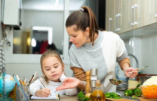 Little girl writing school lesson during mother cooking at kitchen