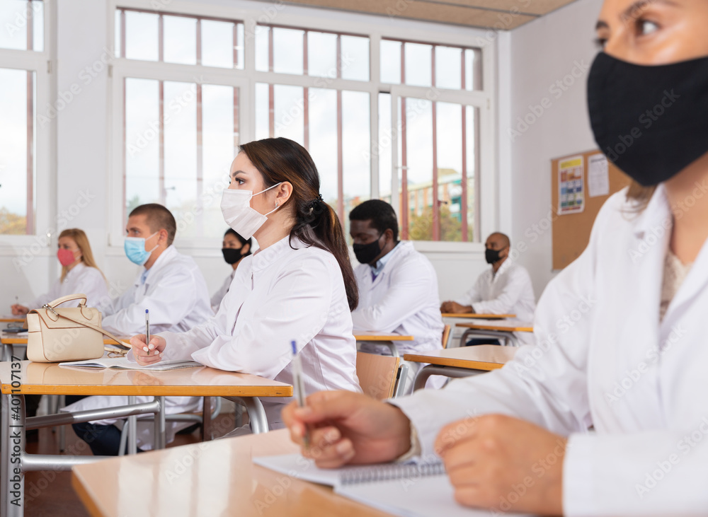 Fototapeta premium Interested female medical student wearing protective face mask making notes during group lecture in classroom. Necessary precautions in public place during coronavirus pandemic