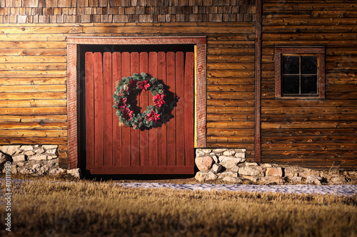 A Christmas wreath hangs on a wood barn door. There is a little snow on the ground. There is also a window off to the right and some stonework at the bottom of the barn. There is wood texture.