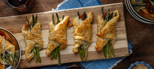 Top down view of asparagus and prosciutto puff pastry bundles on a wooden cutting board with sunlight streaming in.