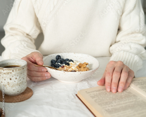 Reading book and eating healthy holiday winter breakfast with granola muesli and yogurt in bowl on white table background. Organic morning diet meal with oat