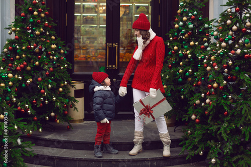 mom in a knitted red sweater and hat stands with the child at the Christmas tree on the street