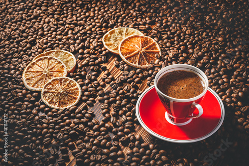 Roasted coffee beans are scattered on the table. Espresso in a red cup on a red saucer. A few slices of dried oranges on coffee beans. © Andrey Ligru