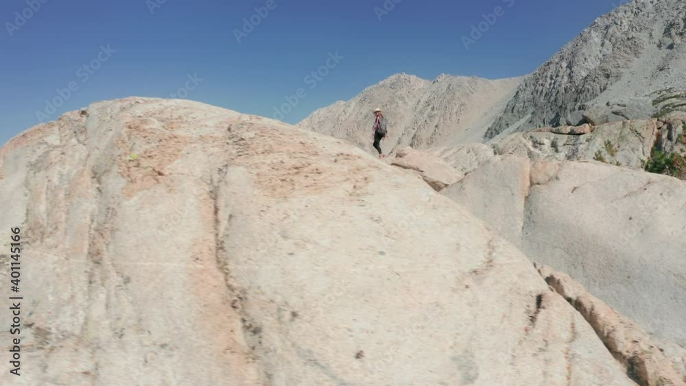 Traveler hiking rocky mountains. Brave young woman exploring the wild nature in rocky mountains on sunny summer or spring day with blue sky above high peaks. Drone flying behind the traveler, 4K USA Traveler hiking rocky mountains. Brave young woman exploring the wild nature in rocky mountains on sunny summer or spring day with blue sky above high peaks. Drone flying behind the traveler, 4K USA