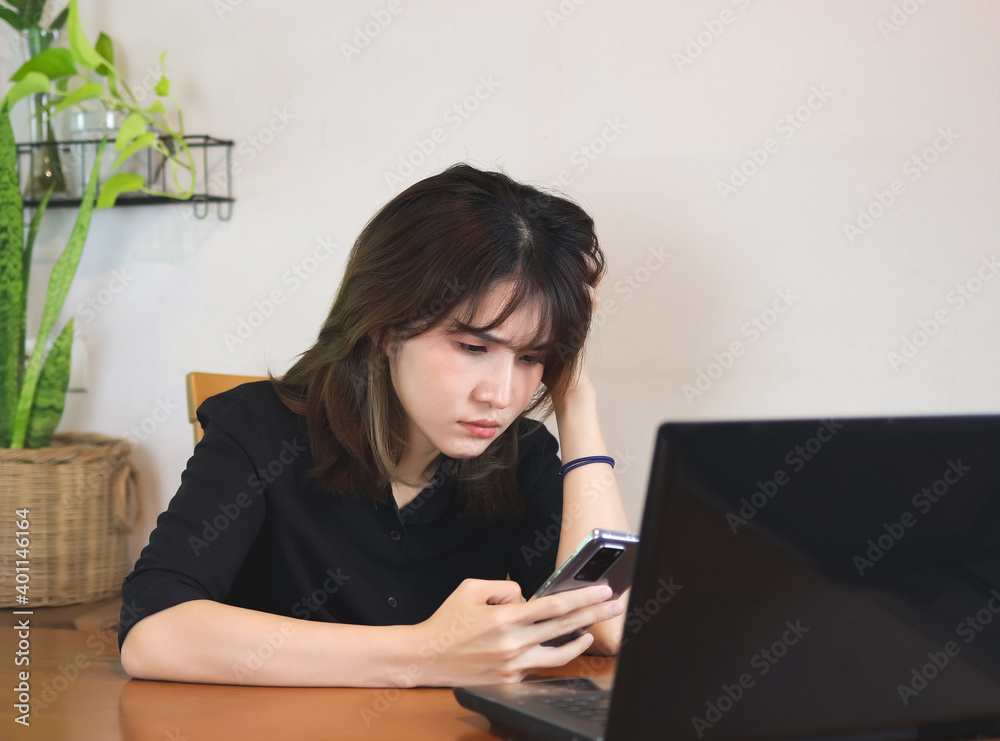 upset Asian woman wearing black shirt, sitting at wooden table with computer laptop, reading message in mobile phone, touching head, feeling worried. white wall with house plant background. upset Asian woman wearing black shirt, sitting at wooden table with computer laptop, reading message in mobile phone, touching head, feeling worried. white wall with house plant background.