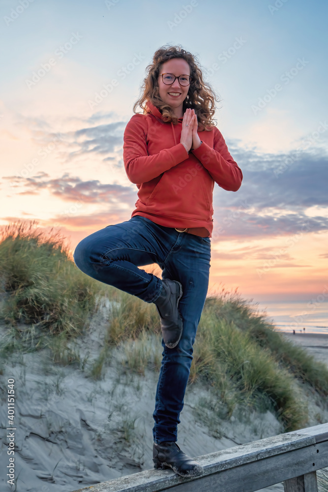 A smiling young woman in casual wear on boots, does a yoga pose on a fence by the beach A smiling young woman in casual wear on boots, does a yoga pose on a fence by the beach