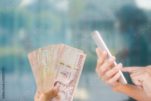 Close up using mobile with thai banknotes 1000 baht on blue background. for accompanying articles on financial transactions through the network banking system. 2021 year Spending money concept.