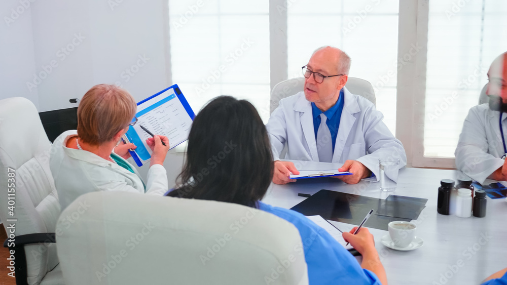 Obraz premium Mature woman doctor pointing on clipboard during briefing with coworkers working in hospital meeting room. Clinic expert therapist talking with colleagues about disease, medicine professional