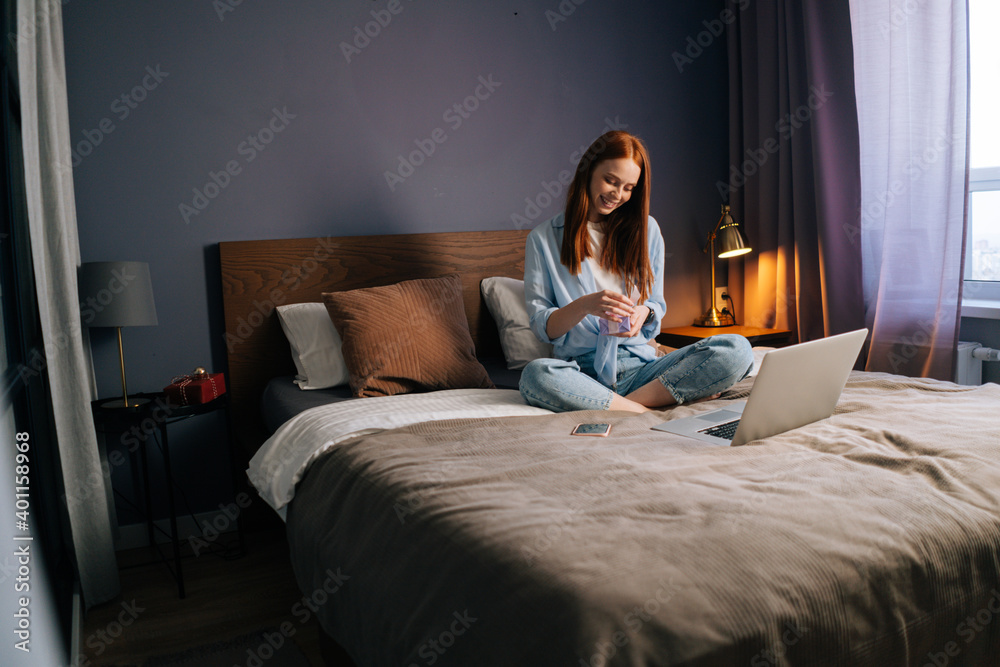 Happy red-haired young woman opening gift box with present at cozy living room and looking on display screen. Cute female having online video chat sitting at bed in light bedroom. Happy red-haired young woman opening gift box with present at cozy living room and looking on display screen. Cute female having online video chat sitting at bed in light bedroom.