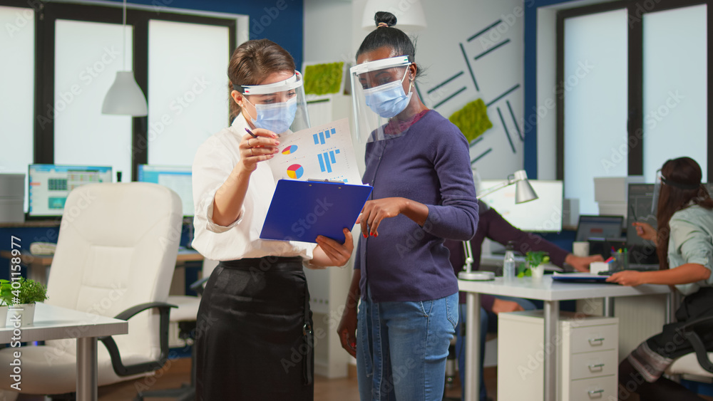 Obraz premium Company manager with visor against coronavirus explaining to african colleague project plan standing in front of desk holding clipboard. Multiethnic business team working respecting social distance.