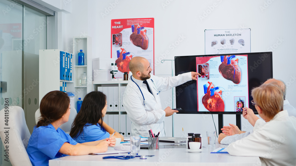 Experienced man doctor analysing heart issues image together with cvalified colleagues in meeting room, pointing on monitor. Doctors discussing diagnosis about treatment of patients Experienced man doctor analysing heart issues image together with cvalified colleagues in meeting room, pointing on monitor. Doctors discussing diagnosis about treatment of patients