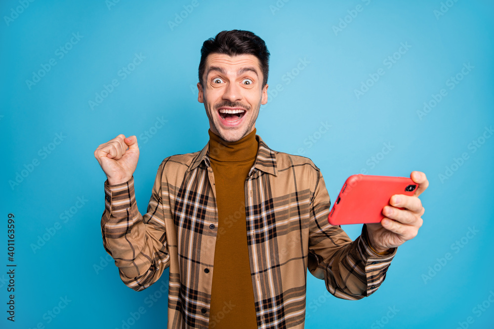 Photo of young happy excited crazy positive man playing video game in phone hold fist in victory isolated on blue color background Photo of young happy excited crazy positive man playing video game in phone hold fist in victory isolated on blue color background