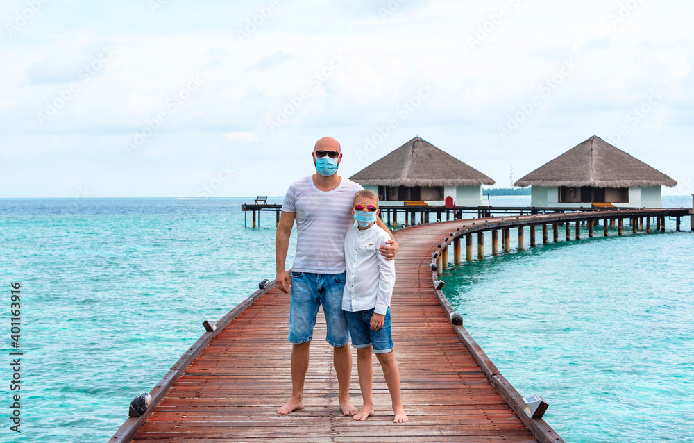 family father young man and his son wearing a protective face mask and stand on a pier near water villas in the Maldives, a concept of  travel during the covid pandemic