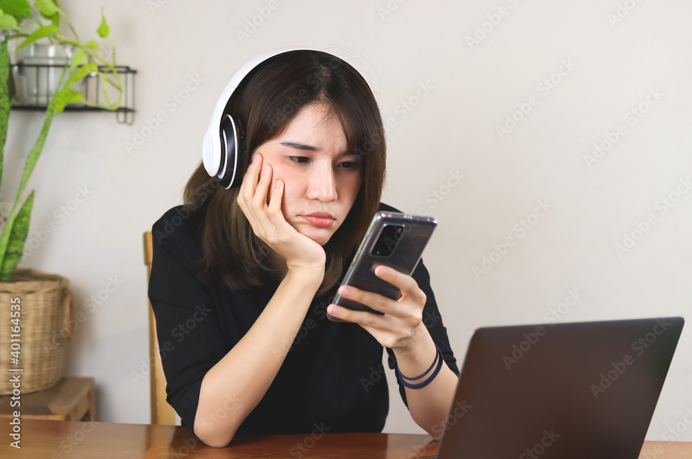 Asian woman wearing headphones, sitting at wooden table with computer laptop, looking seriously at mobile phone, chin on hand. white wall with house plant background. Asian woman wearing headphones, sitting at wooden table with computer laptop, looking seriously at mobile phone, chin on hand. white wall with house plant background.