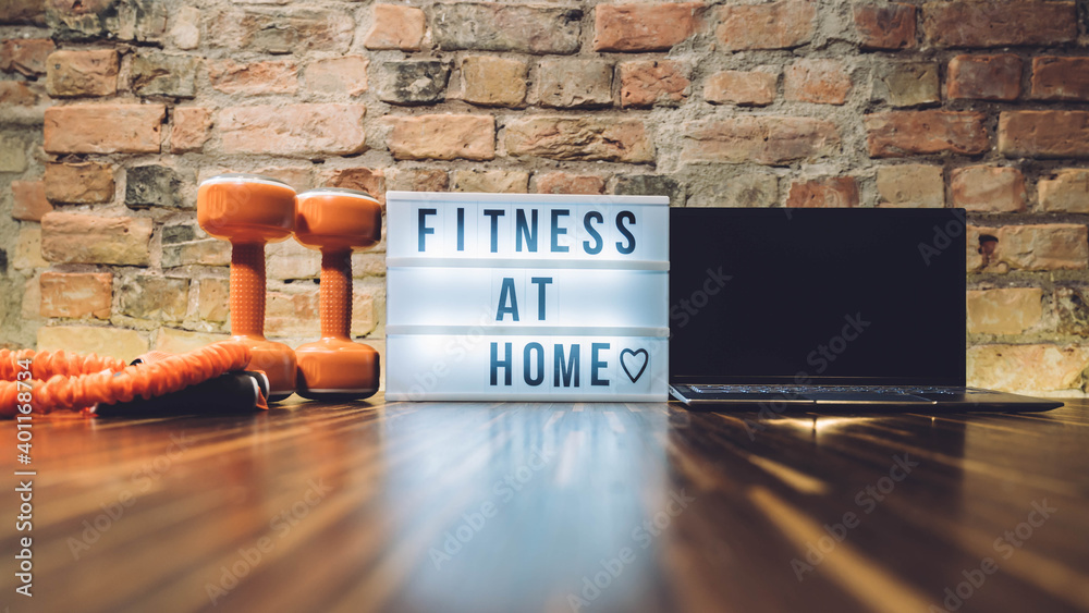 Sport equipment and lightbox with text FITNESS AT HOME on floor indoors. Message to promote self-isolation during COVID‑19 pandemic. Working out at home. Coronavirus COVID-19 Social distancing sign 