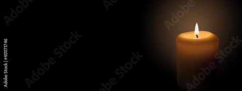 White candle on a black background. The candle flame illuminates the space around it. Banner with space for text.