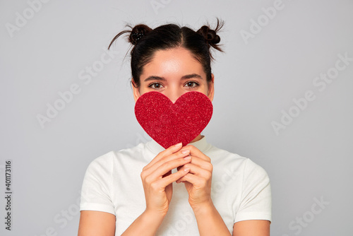 young attractive cheerful ethnicity woman in love holding a big red sparkling heart valentine and covering her mouth and nose while smiling with her eyes