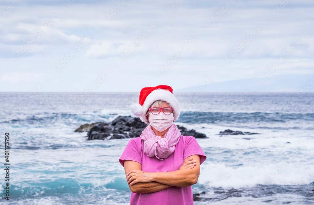 Obraz premium Senior woman in Santa hat wears a surgical mask due to coronavirus infection and enjoys the winter sea looking at camera. Horizon over water behind her