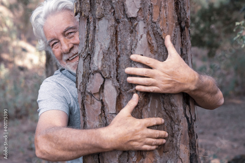 Valokuva An adult man with a beard and white hair smiles as he hugs a tree trunk in the woods