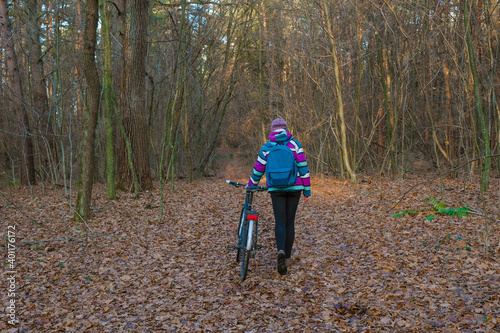 A woman with a bicycle on a forest walk in the cold season. Bright clothes on a blurred brown forest background. Copy space. 
