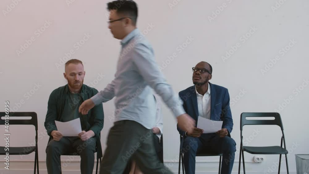 Medium long of three nervous multiethnic job applicants sitting on chairs in hallway, waiting for their turn, angry man walking by, unrecognizable manager inviting Caucasian man for interview Medium long of three nervous multiethnic job applicants sitting on chairs in hallway, waiting for their turn, angry man walking by, unrecognizable manager inviting Caucasian man for interview