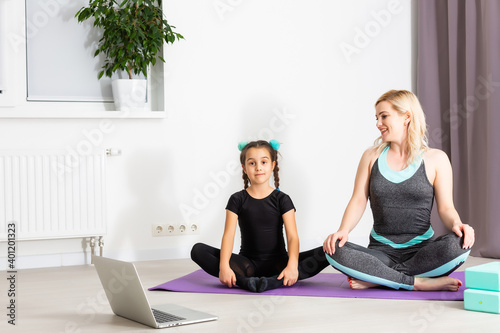 Mom and little daughter are doing gymnastics on the mat at home. They do yoga. They are fun because they have a happy family. Poses they are looking at the laptop.