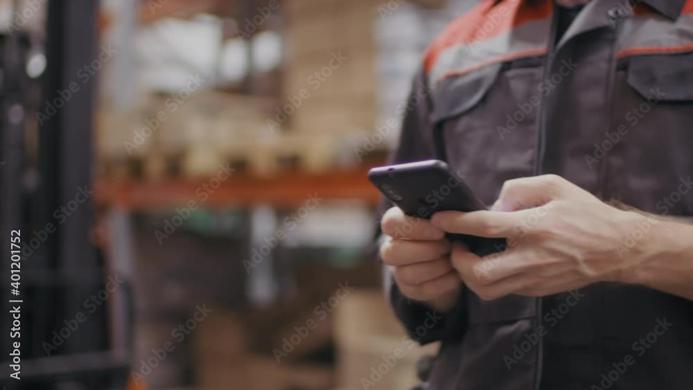 Midsection slow-motion shot of unrecognizable hands of workman in construction overall suit holding smartphone texting messages while walking at warehouse