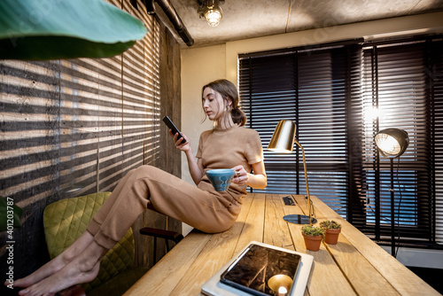 Relaxed woman in comfortable domestic suit with a smart phone and coffee cup sitting on the table at the beautiful home office