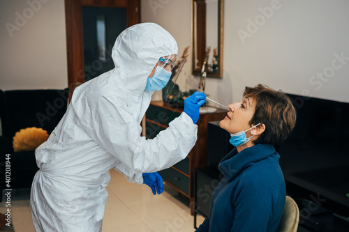 Health Professional in PPE, face mask and shield, introducing a nasal swab to a senior female patient on a home visit. Rapid Antigen Test to analyze nasal sampling for Covid-19, Coronavirus Pandemic.