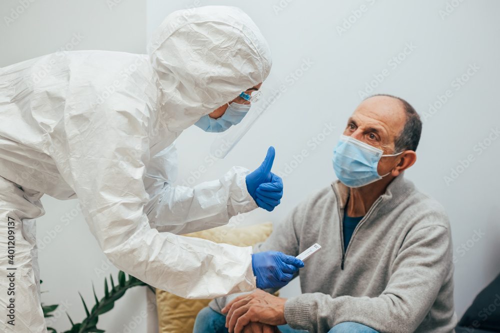 Fototapeta premium Health professional in a PPE suit, mask, face shield showing the patient the negative result for Covid-19. Antigen test to an elderly patient during a home visit. Coronavirus Pandemic.